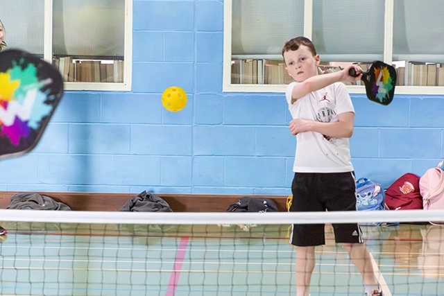 Children playing pickleball indoors with a yellow ball.