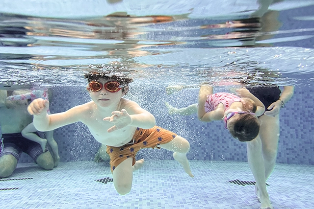 Two toddlers swim underwater in the learner pool at West Wight Sports and Community Centre. Two adults can be partially seen in the background.