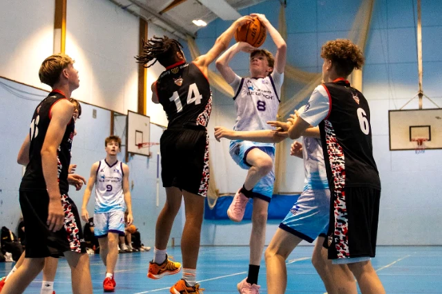 A group of young people play basketball ina sports hall. Two players in the middle have jumped. One tries to stop the opther from shooting a basket.
