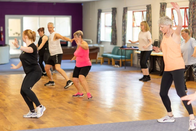 An instructor leads a class of Dance Fit. Attendees in colourful gym wear reach upward mid-move.