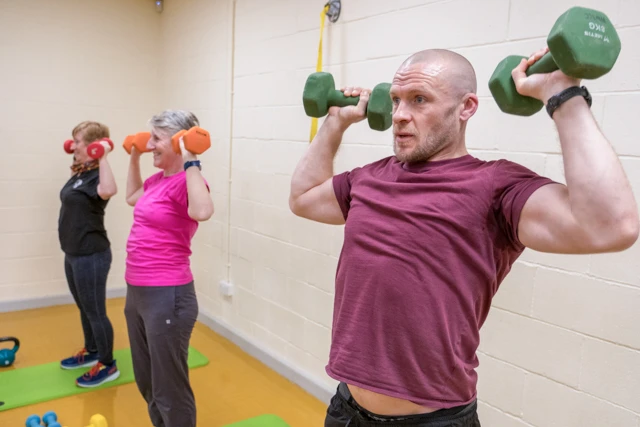 Three people are exercising with dumbbells in a gym. A man in the foreground is performing an overhead press with green dumbbells, while two women in the background are doing the same exercise with orange and red dumbbells respectively.