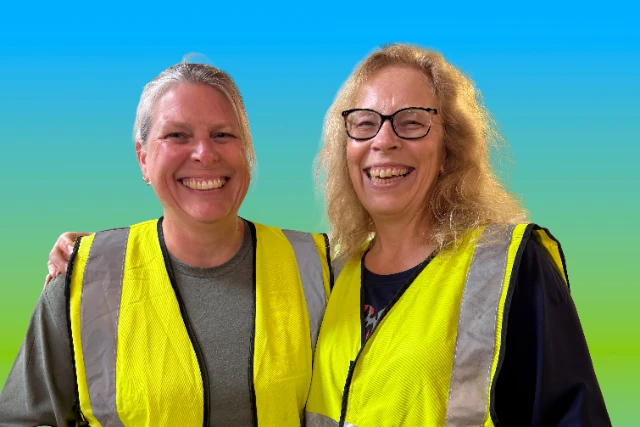Marshalls (1) Two women wearing high-visibility vests are smiling and posing together volunteering at an event for West Wight Sports and Community Centre. The background is a gradient of blue and green.