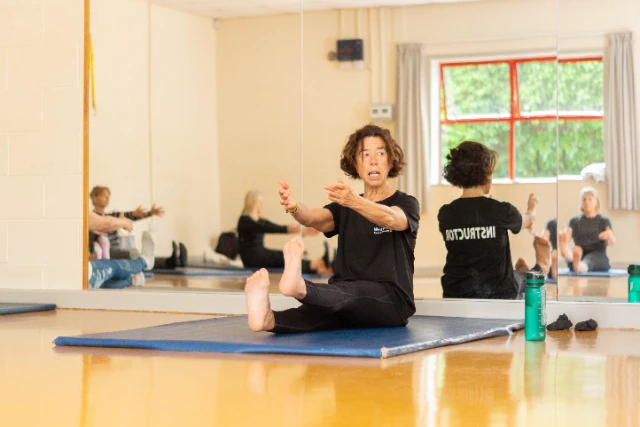 Female class leader demonstrates a move within the tone and stretch class. Attendees can be seen replicating this move in the mirror.