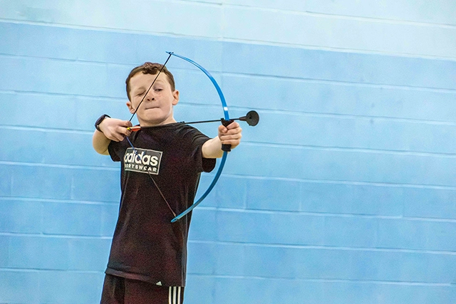 A young boy in a black t-shirt draws back a blue bow, aiming forward. He is practicing archery in front of a light blue wall.