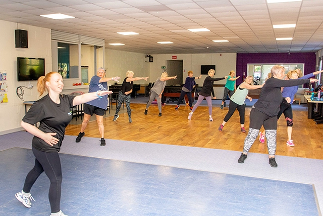 A group of women, led by an instructor in the foreground, are participating in an exercise class in a community hall. They are all in workout attire and performing similar arm movements.