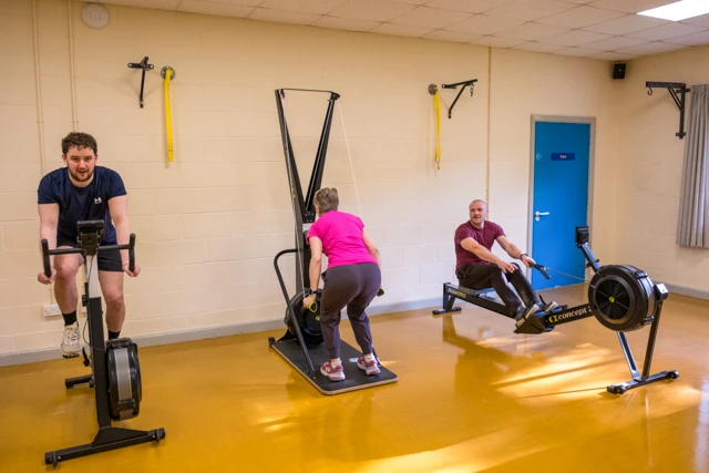 Three people are exercising during HIIT class using different machines. A man is on a stationary bike, a woman is on a ski machine, and another man is on a rowing machine.