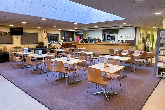 This image shows an empty cafe dining area with tables and chairs. A counter with kitchen equipment is visible in the background.