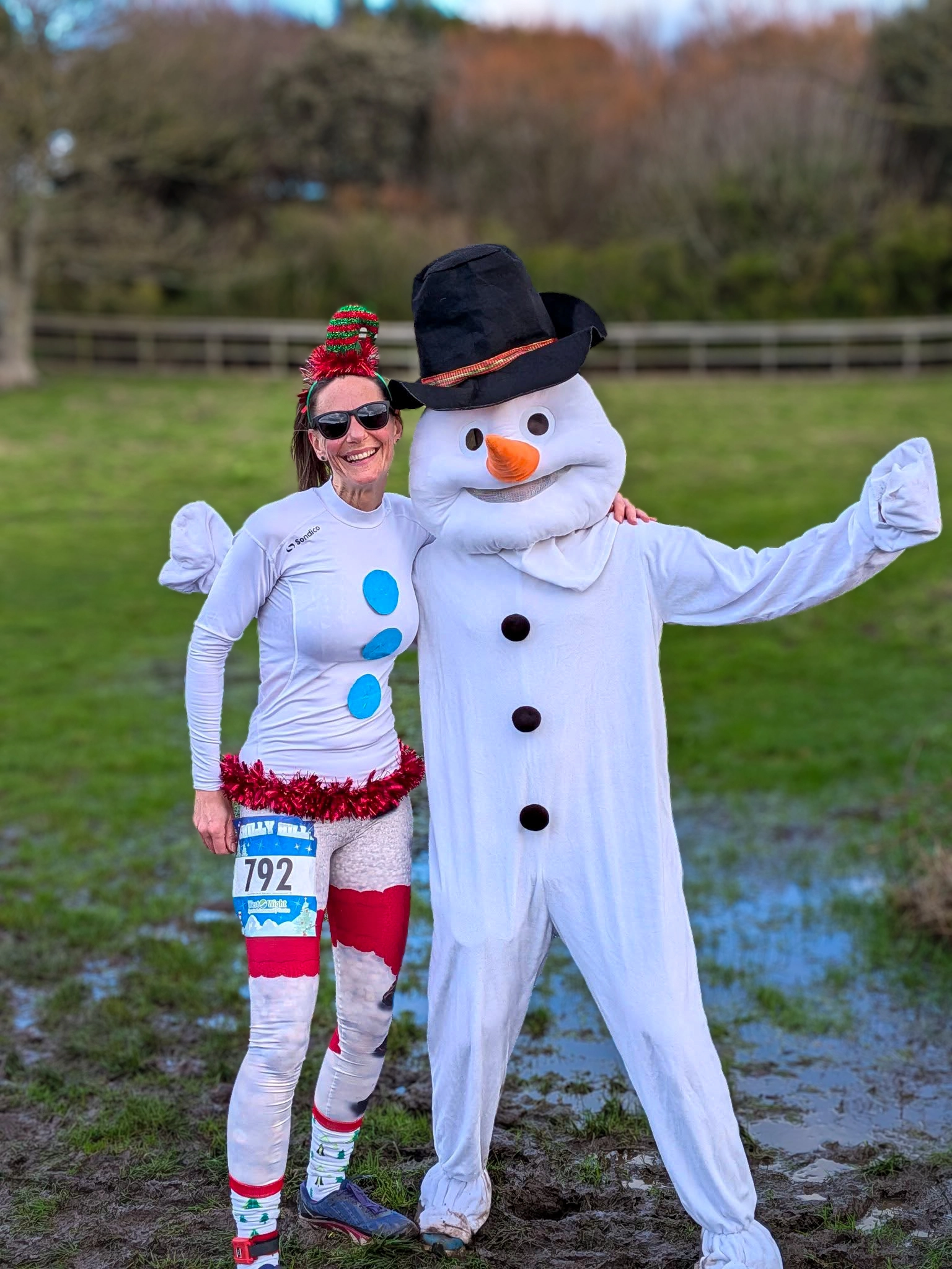 A woman dressed in a festive snowman costume poses with a person in a full snowman mascot suit. They are standing outdoors in a grassy area with trees in the background.