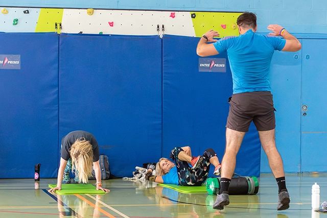 An instructor in a blue shirt and shorts stands facing away from the camera, leading a circuits class. Two women are on the floor on mats, one in a plank position and the other lying on her back.