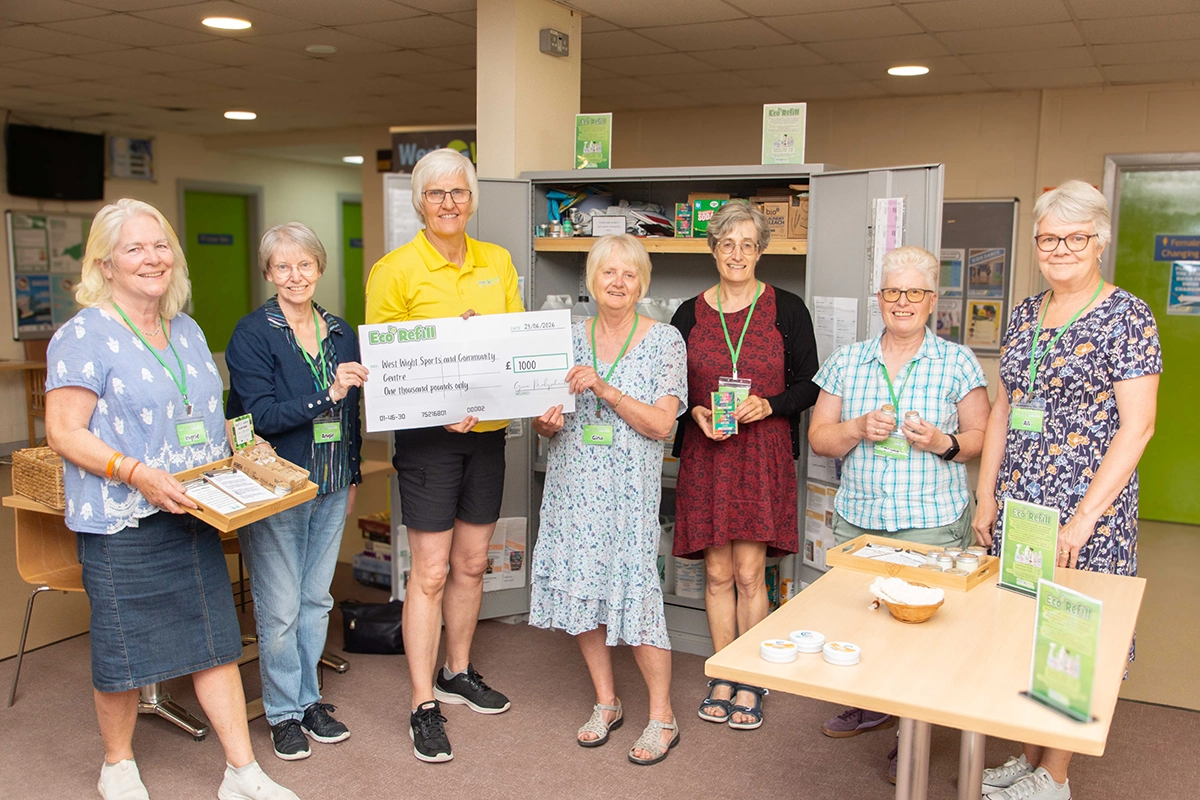 A group of female Eco Refill volunteers are posing for a photo, alongside centre manager, Clare Griffin. They are presenting a £1000 cheque. Several women are wearing lanyards with name tags. There are also items displayed on a table in the foreground, related to the eco-refill service.
