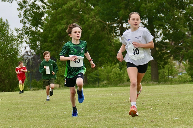 Fun Run 2026 Children are running a race on a grassy field with trees in the background. The two children in the foreground are wearing running bibs