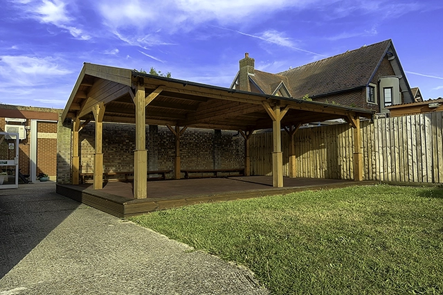 The Garden Canopy, a wooden outdoor shelter with a raised deck stands in a yard next to a brick building and a wooden fence. The sky above is blue with wispy white clouds.