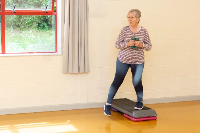 An elderly woman is exercising with hand weights while standing on a step aerobics platform. She is wearing a striped long-sleeved shirt and ombre leggings.