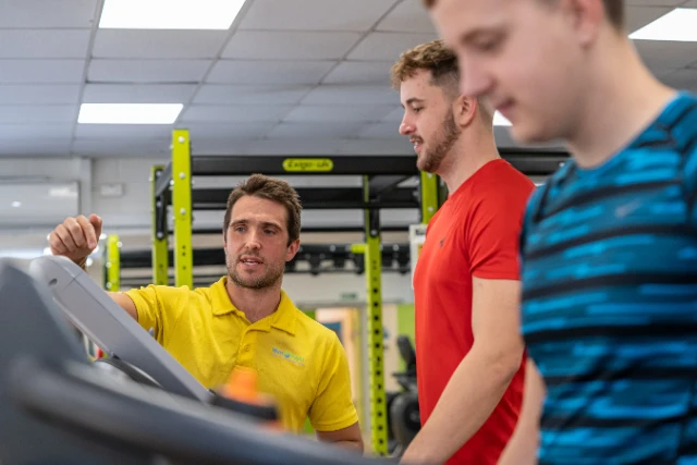 Gym (68) A fitness instructor in a yellow shirt points to a treadmill while two other men stand nearby in a gym.