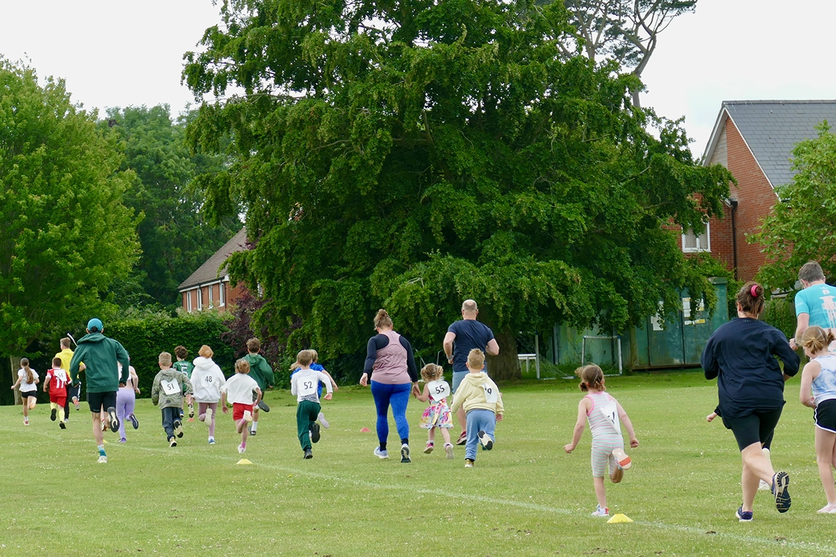 A group of children and adults are participating in a race on a grassy field. They are running away from the camera, with trees and houses in the background.