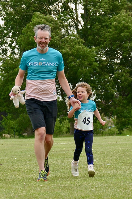 Junior fun run 001 A man holding a teddy bear and a young girl are running together on a grassy field. They are both wearing matching shirts.