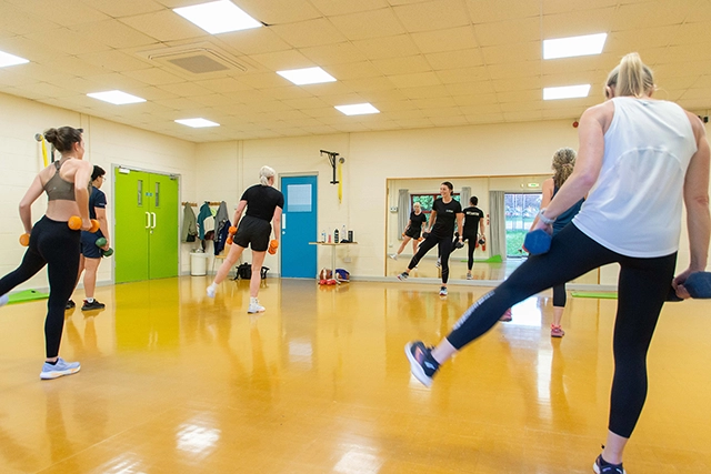 A group of women are participating in a fitness class, performing exercises with dumbbells. They are in a brightly lit room with a shiny yellow floor and mirrors on one wall.