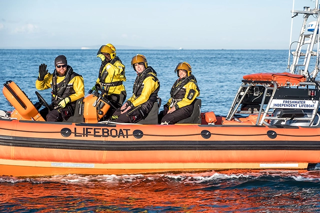 Lifeboat Four individuals in yellow and black safety gear are on an orange lifeboat in the water. The lifeboat has "LIFEBOAT" printed on its side and a sign that reads "FRESHWATER INDEPENDENT LIFEBOAT".