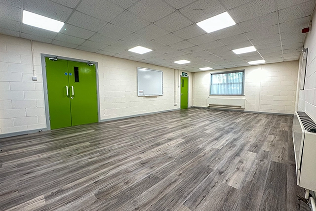 This is an interior view of an empty meeting room with grey wood-look flooring. The room features white brick walls, fluorescent lighting, and two bright green doors.