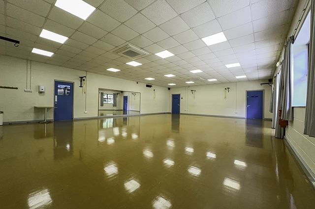 An empty dance studio with a polished floor and mirrors on the wall. There are two blue doors visible and large windows with curtains.