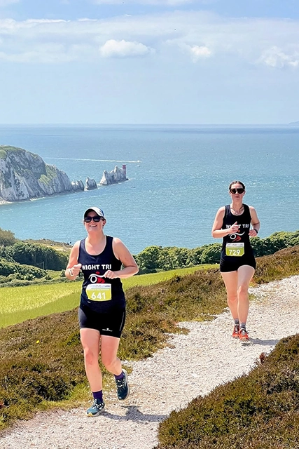 Two women are running on a scenic coastal trail during a triathlon race. The background features the iconic Needles coastline and the ocean under a partly cloudy sky.