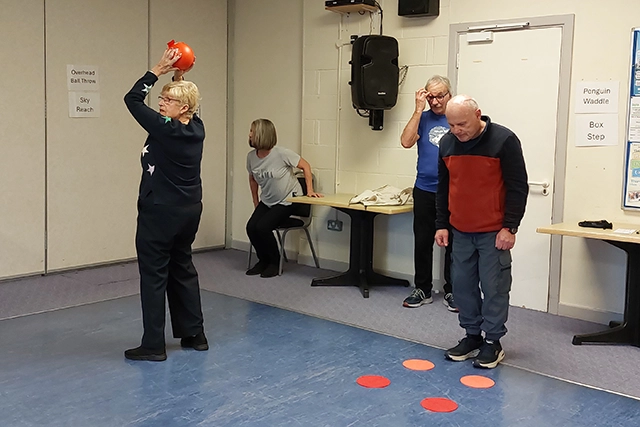 Parkinson's course 640 A group of adults are participating in a Parkinson's exercise class in a room. A woman in the foreground is holding an orange ball above her head, for an overhead ball throw exercise.