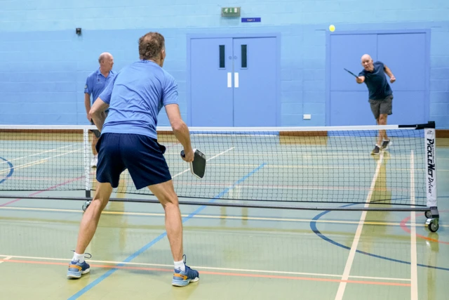 Three men play pickleball indoors on a court. One man is hitting the ball, while two others are positioned on the other side of the net.
