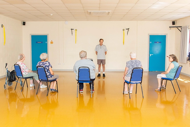 Really Useful cafe A group of older adults are seated in chairs facing a male instructor in a room with a bright yellow floor. The instructor stands in front of them, and they appear to be participating in the Reall Useful Cafe session.