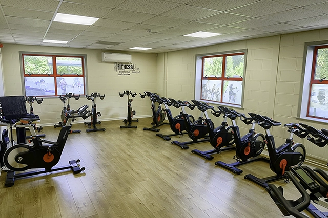 An indoor cycling studio is filled with rows of black and red stationary bikes. The room has a light-colored wooden floor and beige walls with windows showing outdoor scenery and motivational text.