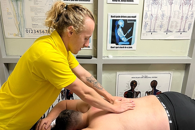 Soft Tissue Therapy A therapist in a yellow shirt gives a massage to a person lying on their stomach. Anatomical charts are visible in the background.