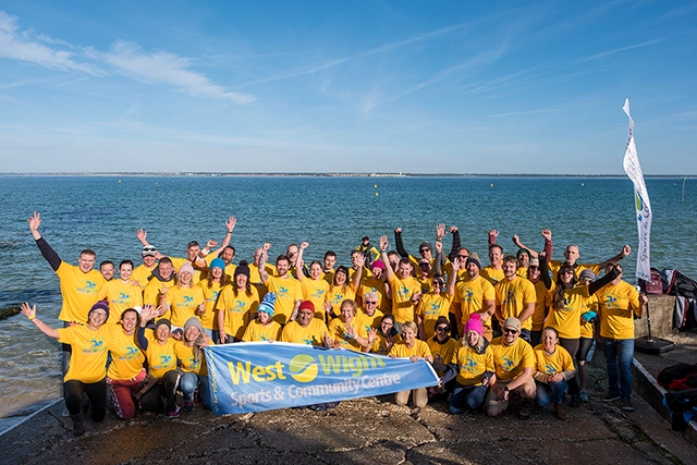 A large group of people wearing yellow shirts and holding a West Wight Sports and Community Centre banner on Colwell shoreline after completing the Solent Swim
