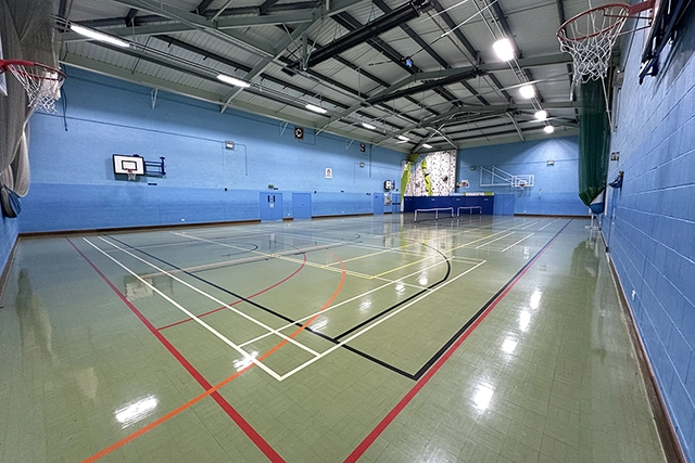 Sports Hall An indoor sports hall with a basketball court, tennis court, and a climbing wall is shown. The room is brightly lit with fluorescent lights.