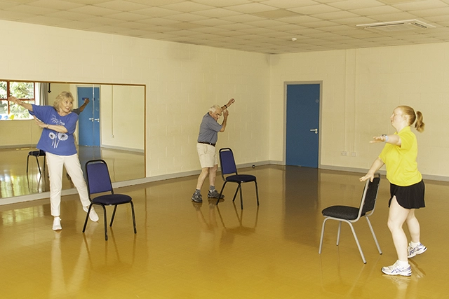 Two people are participating in a Strength and Balance exercise class in a room with a mirrored wall. They are all in motion, with one woman in blue and an older man in a blue shirt and shorts, led by a female instructor in a yellow shirt.
