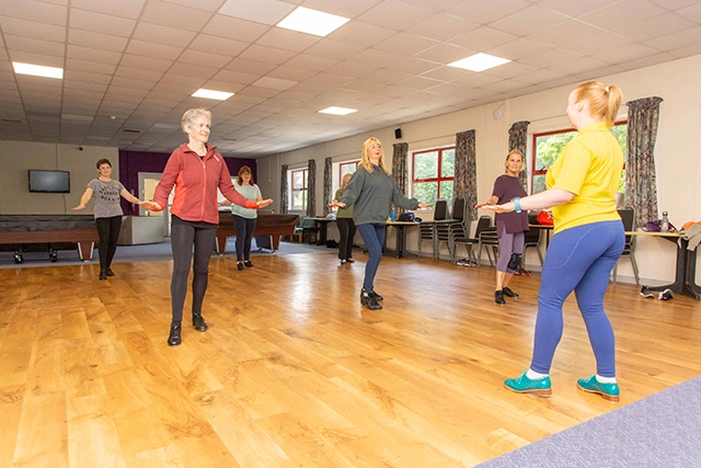 A group of women are participating in a tap dance class in the community hall. They are all standing and following along with an instructor who is facing away from the camera.