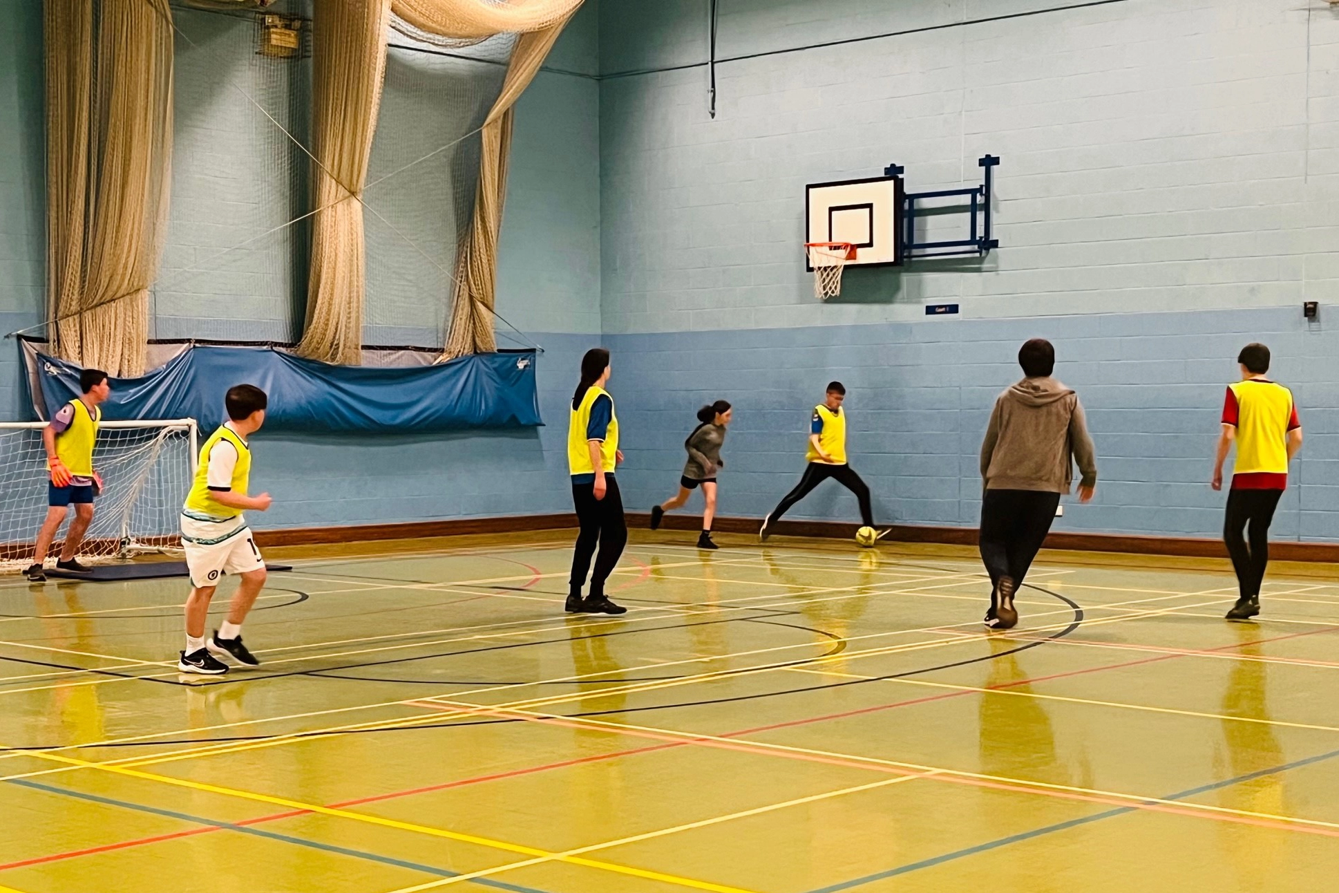 A group of young people are playing football in the sports hall. They are wearing brightly colored vests over their athletic clothing.